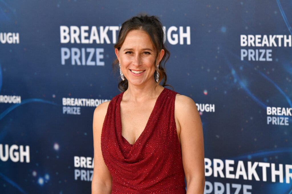 Anne Wojcicki mit einem roten, tief ausgeschnittenem Kleid vor einem Plakat mit der Aufschrift: Breakthrough Prize