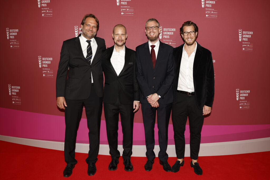 Jonas Andrulis mit vier weiteren Männern auf der Bühne die einen roten Hintergrund und der Beschriftung Deutscher Gründer Preis zeigt