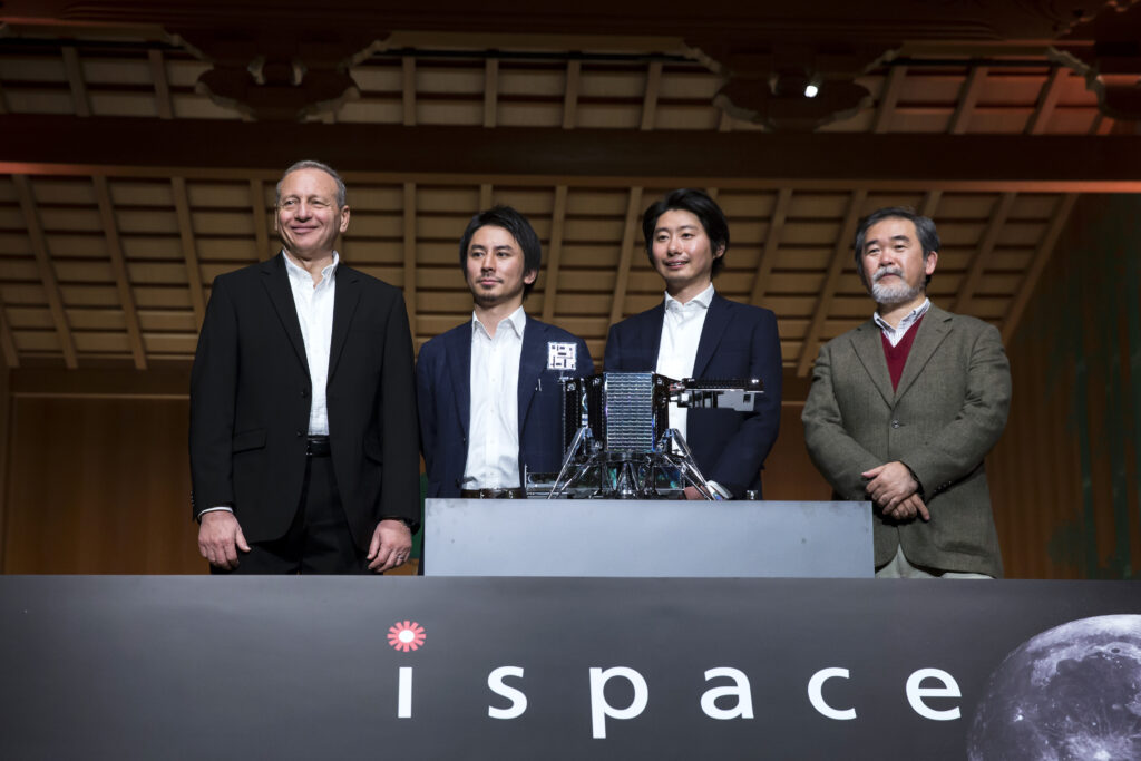 Takeshi Hakamada steht neben drei anderen Männern auf einer Bühne. Vor ihnen ein Modell eines Fahrzeuges, das für die Mondbefahrung geplant ist
