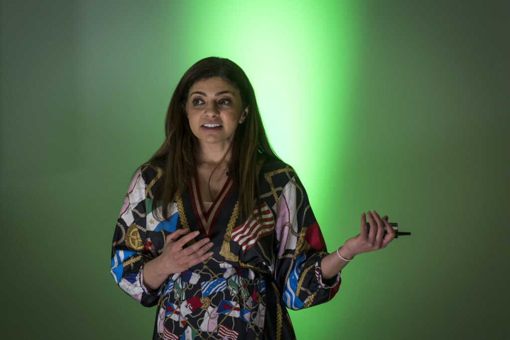 Dr Rana el Kaliouby mit einer bunten Bluse vor einem grünen Licht. Sie hält einen Beamer in der einen Hand und spricht zu Menschen