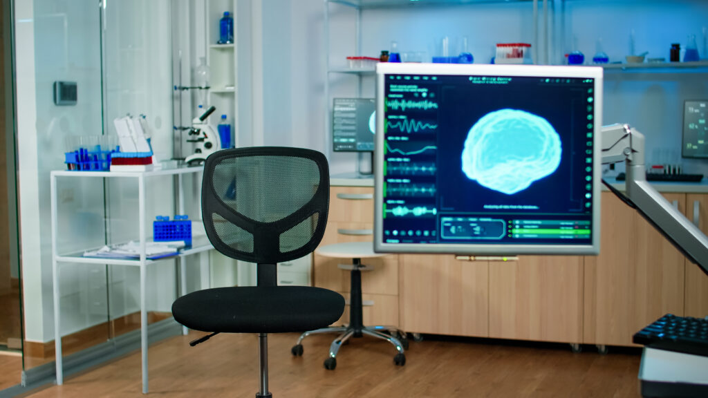 Blick in ein Labor eines Neurowissenschafters. Ein schwarzer Stuhl, ein Mikroskoph sowie ein Monitor dass das menschliche Gehirn zeigt