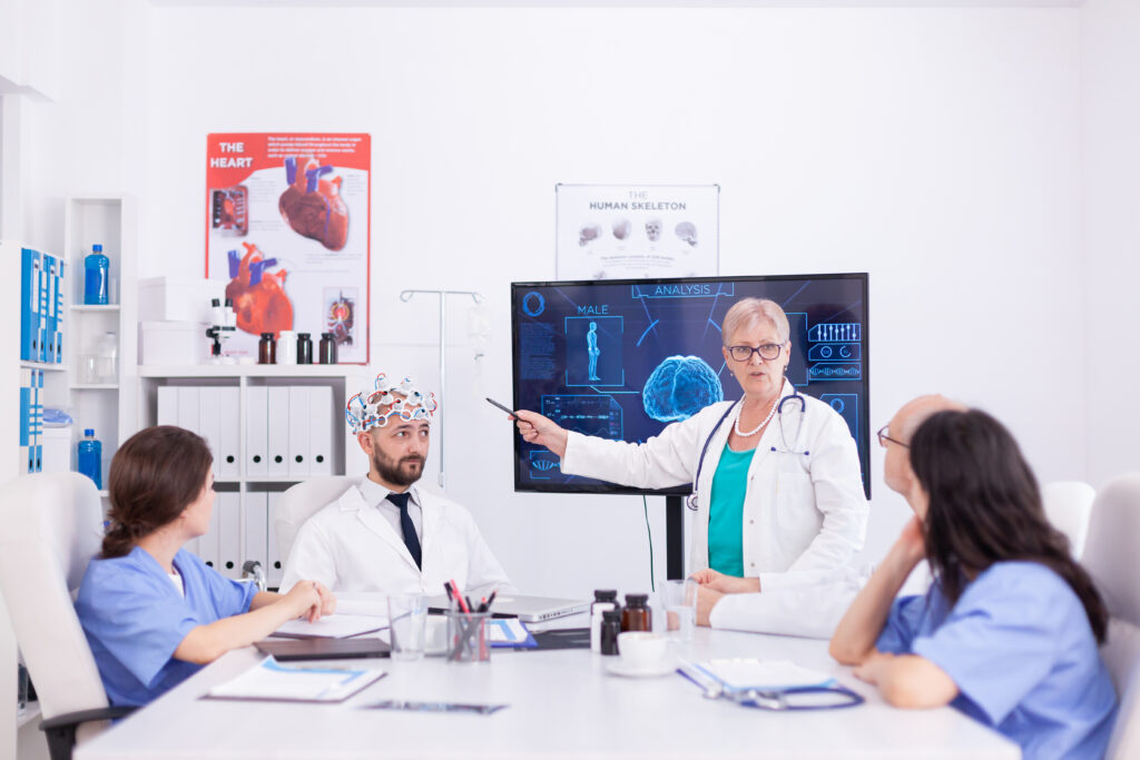 Eine Ärztin trägt einen weißen Mantel und ein türkises T-Shirt. Sie steht vor einem Monitor und zeigt mit einem Stift au ein Bild. Vor hier sitzen vier weitere Kollegen. Ein Mann trägt eine Kappe die Gehirnverbindungen darstellen