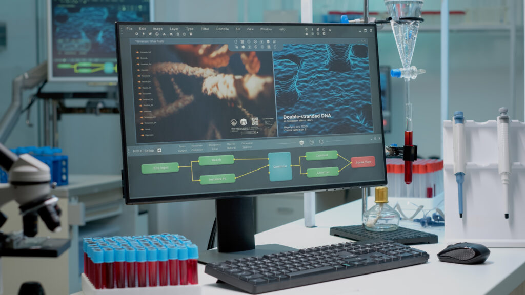 Blick auf einen Computer in einem Labor. Davor eine Tastatur, zwei Pipetten und eine Menge an Pipetten mit blauen Verschlüssen und roter Flüssigkeit