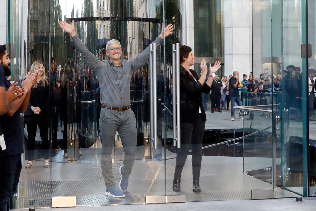 Tim Cook mit grauer Anzughose und grauem Hemd tritt vor viele Menschen und reißt dabei die Hände in die Höhe