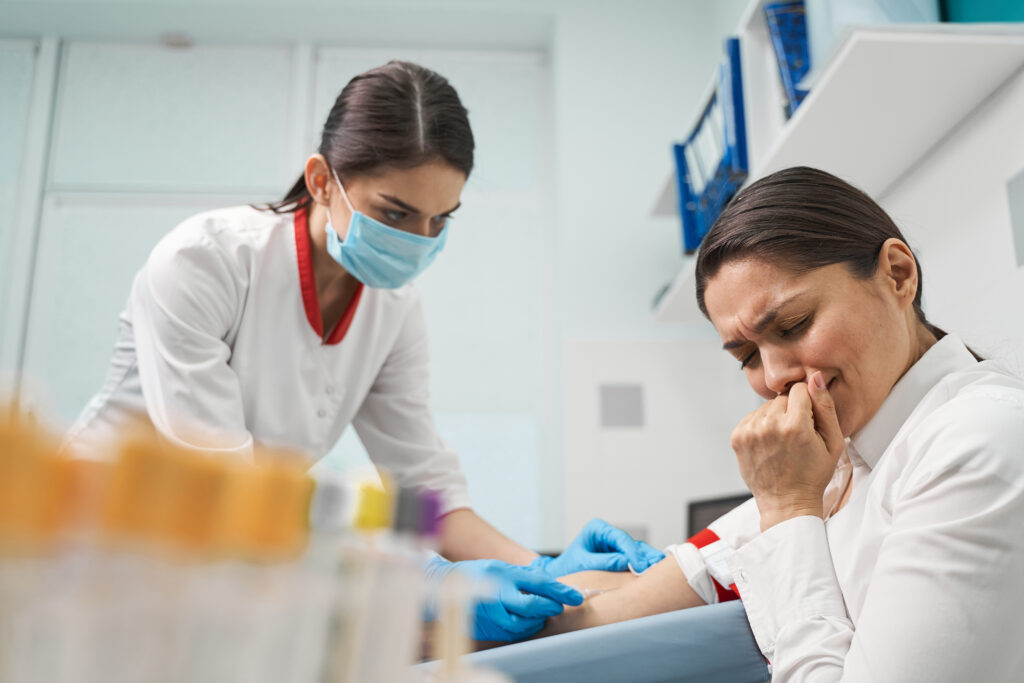 Frau mit Mundschutz und weißer Kleidung hält die Hand einer Patientin die das Gesicht vor Schmerzen verzieht. 