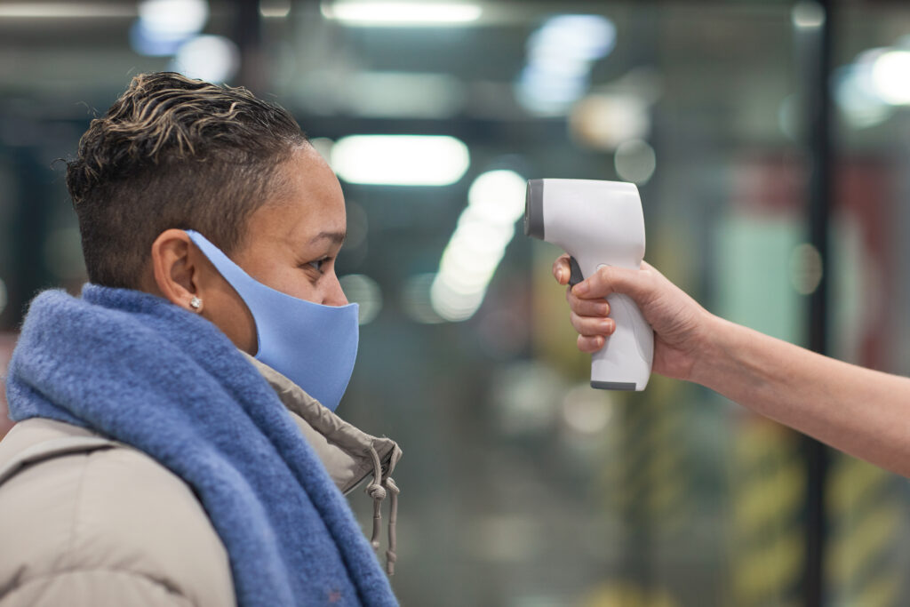 Frau mit blauem Mundschutz und vor ihr eine Hand mit einem großen Fieberthermometer