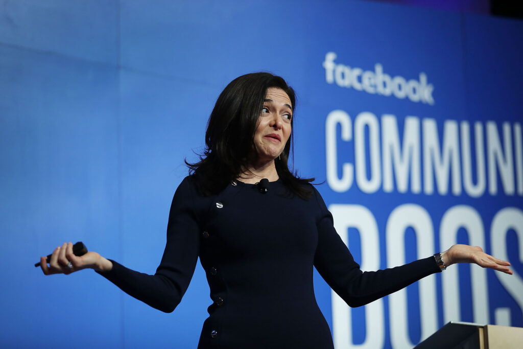 Sheryl Sandberg steht auf einer Bühne und hält die Hände weit geöffnet. Hinter ihr eine Leinwand von Facebook